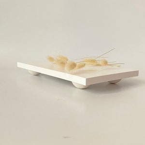 Puede incluir: Bandeja rectangular blanca con bordes redondeados y cuatro pequeños pies blancos. La bandeja está vacía y tiene una rama de flores blancas secas en la parte superior.