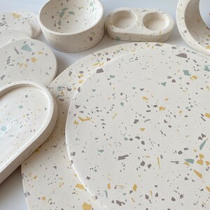 Pode incluir: Um conjunto de  copos de apoio, tigelas e bandejas de terrazzo branco em v&aacute;rios tamanhos. Os copos de apoio e as bandejas apresentam um padr&atilde;o salpicado com manchas amarelas, verdes e marrons.