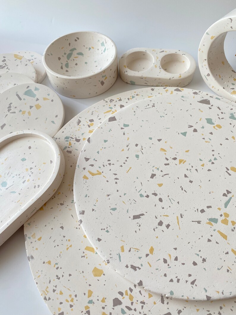 Pode incluir: Um conjunto de  copos de apoio, tigelas e bandejas de terrazzo branco em v&aacute;rios tamanhos. Os copos de apoio e as bandejas apresentam um padr&atilde;o salpicado com manchas amarelas, verdes e marrons.