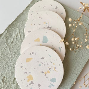 Pode incluir: Quatro  copos de apoio redondos brancos com um padr&atilde;o de terrazzo em tons de azul claro, amarelo e cinza. Os copos de apoio est&atilde;o empilhados um em cima do outro.