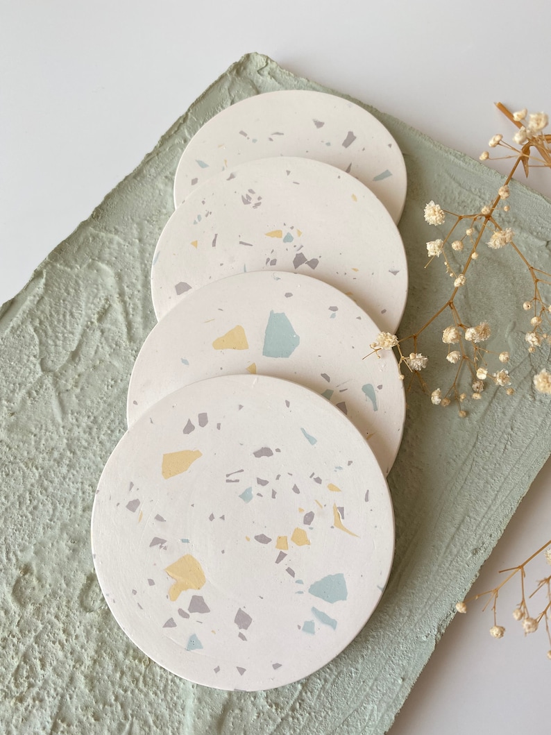 Pode incluir: Quatro  copos de apoio redondos brancos com um padr&atilde;o de terrazzo em tons de azul claro, amarelo e cinza. Os copos de apoio est&atilde;o empilhados um em cima do outro.