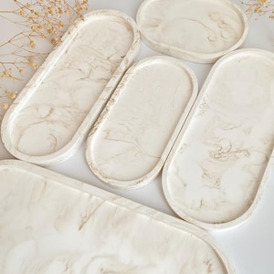 以下が含まれることがあります： 白い大理石模様の仕上げが施された、楕円形のトレイ5個セット。トレイは樹脂製で、滑らかで光沢のある表面をしています。