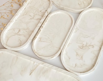Puede incluir: Conjunto de cinco bandejas ovaladas con un acabado blanco marmoleado. Las bandejas están hechas de resina y tienen una superficie lisa y brillante.