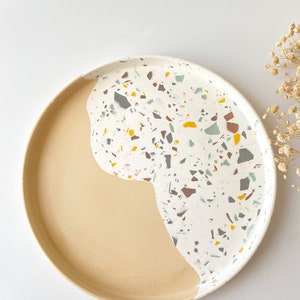 Pode incluir: Uma bandeja redonda de terrazzo bege e branco com um design salpicado. A bandeja é perfeita para servir petiscos, bebidas ou para exibir itens decorativos.