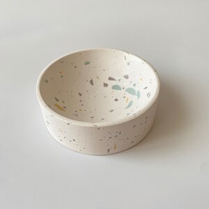 Pode incluir: Uma tigela branca pequena com um design de terrazzo salpicado em tons de cinza, azul e amarelo. A tigela &eacute; redonda e tem uma borda ligeiramente curva.