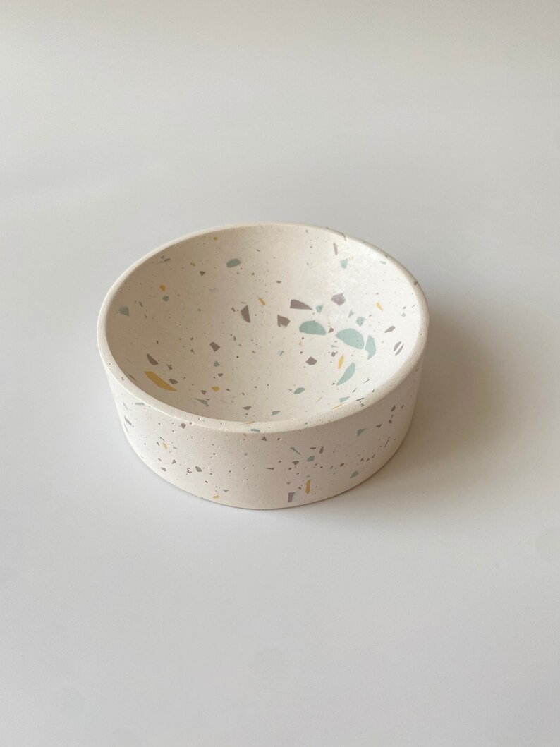 Pode incluir: Uma tigela branca pequena com um design de terrazzo salpicado em tons de cinza, azul e amarelo. A tigela &eacute; redonda e tem uma borda ligeiramente curva.