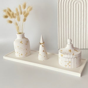 Pode incluir: Bandeja de cerâmica branca com três itens decorativos: um vaso com grama de pampas seca, um porta-anel e uma pequena caixa com tampa. Todos os itens têm uma superfície texturizada e detalhes dourados.