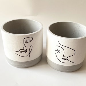 May include: Two gray concrete planters with black line drawings of faces. One planter has a drawing of a face with a closed eye and the other planter has a drawing of a face with an open eye.