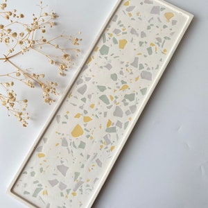 May include: A rectangular white tray with a terrazzo pattern in shades of gray, green, and yellow. The tray is made of resin and has a smooth, polished finish.
