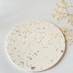 Pode incluir: Uma bandeja redonda de terrazzo branca com um padr&atilde;o salpicado de amarelo, cinza e azul. A bandeja est&aacute; sobre um fundo branco.