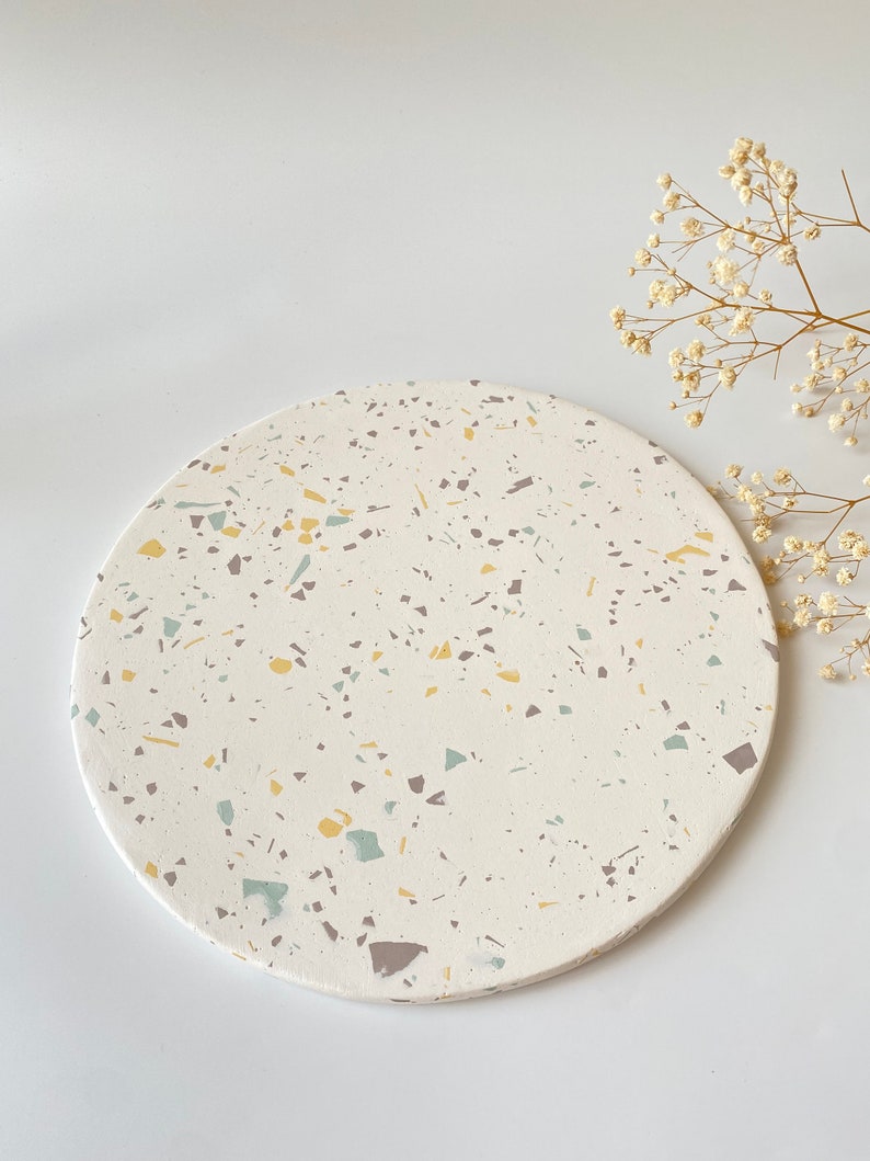 Pode incluir: Uma bandeja redonda de terrazzo branca com um padr&atilde;o salpicado de amarelo, cinza e azul. A bandeja est&aacute; sobre um fundo branco.