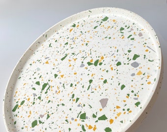 May include: Oval serving tray with a white base and a terrazzo pattern of green, yellow, gray, and purple flecks. The tray has a slightly raised edge and a smooth surface. The tray is suitable for serving food or displaying decorative items.