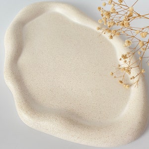 May include: A white, organic-shaped ceramic tray with a speckled texture. The tray is perfect for displaying small items or jewelry.