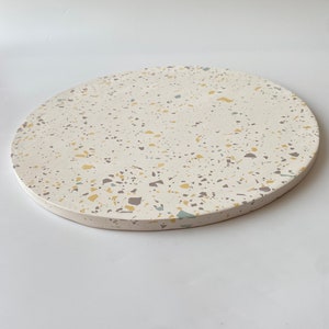 Pode incluir: Uma bandeja redonda de terrazzo branca com um padr&atilde;o salpicado de amarelo, cinza e azul.