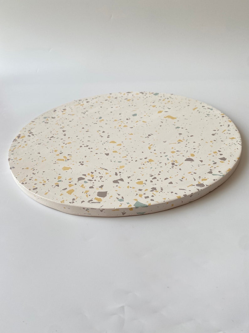 Pode incluir: Uma bandeja redonda de terrazzo branca com um padr&atilde;o salpicado de amarelo, cinza e azul.