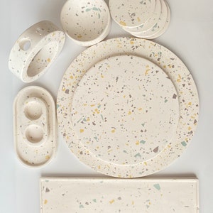 Pode incluir: Um conjunto de pratos de servir de cer&acirc;mica branca com um padr&atilde;o salpicado em tons de amarelo, verde e cinza. O conjunto inclui um prato redondo grande, um prato redondo menor, um prato retangular, um prato oval pequeno, uma tigela redonda e quatro  coasters redondos.