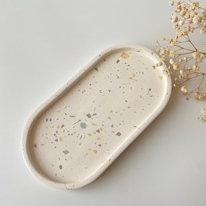 Pode incluir: Uma bandeja oval branca com um design de terrazzo salpicado em tons de bege, amarelo e cinza.