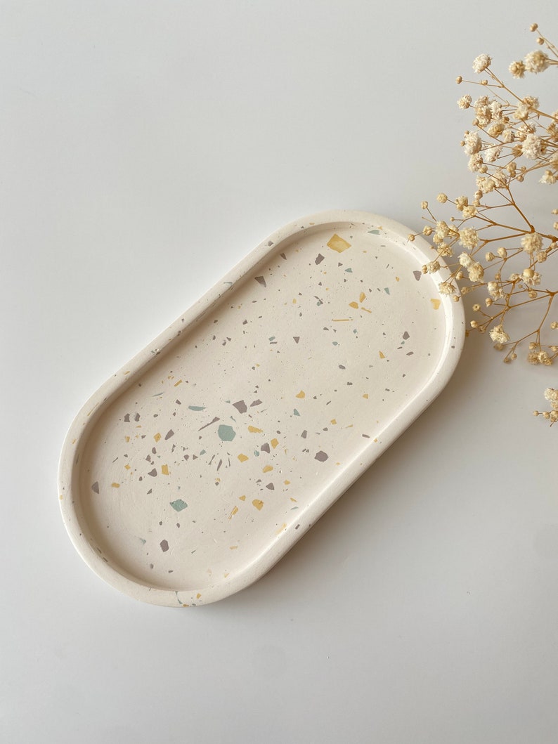 Pode incluir: Uma bandeja oval branca com um design de terrazzo salpicado em tons de bege, amarelo e cinza.
