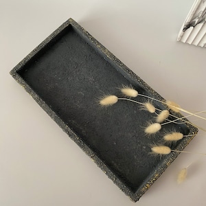 Può includere: Un vassoio rettangolare nero con un bordo punteggiato d'oro. Il vassoio è in cemento e ha una texture ruvida.