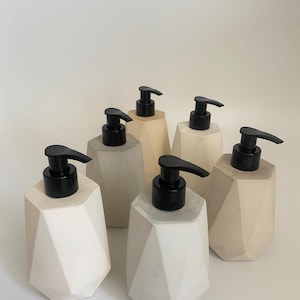 Puede incluir: Cinco dispensadores de jabón de forma geométrica con bombas negras. Los dispensadores están hechos de un material similar al hormigón gris claro. Los dispensadores están dispuestos en fila sobre una superficie blanca.