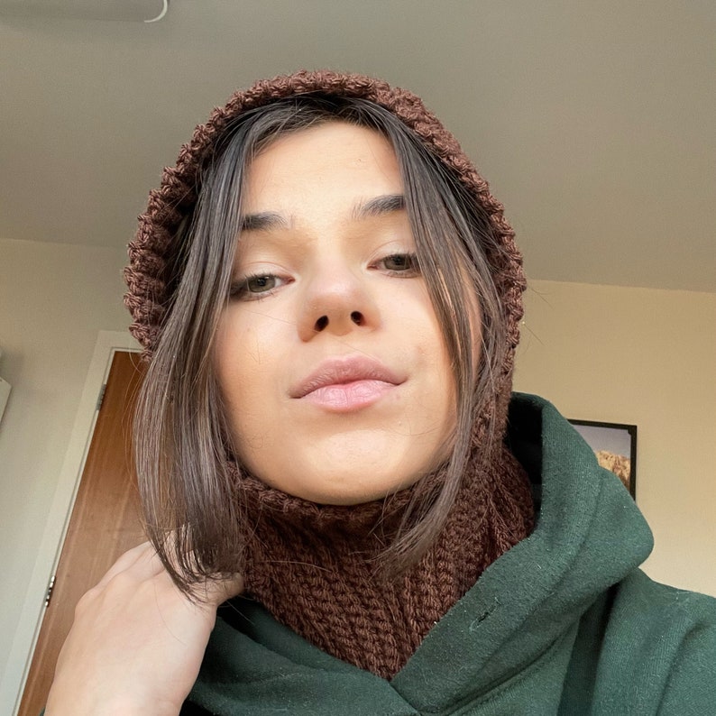 May include: A woman wearing a brown knitted hat and matching neck warmer. She is wearing a dark green hooded sweatshirt.