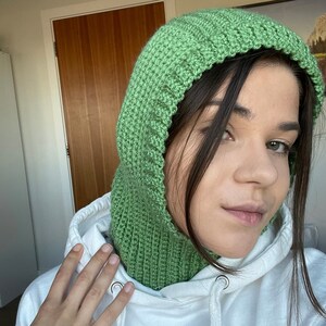 May include: A green crocheted hooded neck warmer. The hood is pulled up over the person's head, and the neck warmer is worn over a white sweatshirt.