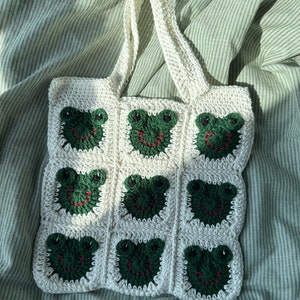 May include: White crocheted tote bag with green frog faces. The bag has a single handle and is made of a soft, textured yarn.