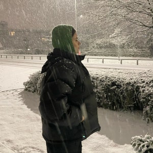 May include: A person wearing a green knitted hat and a black puffer jacket stands in a snowy landscape. The snow is falling heavily and the person is looking off to the side.