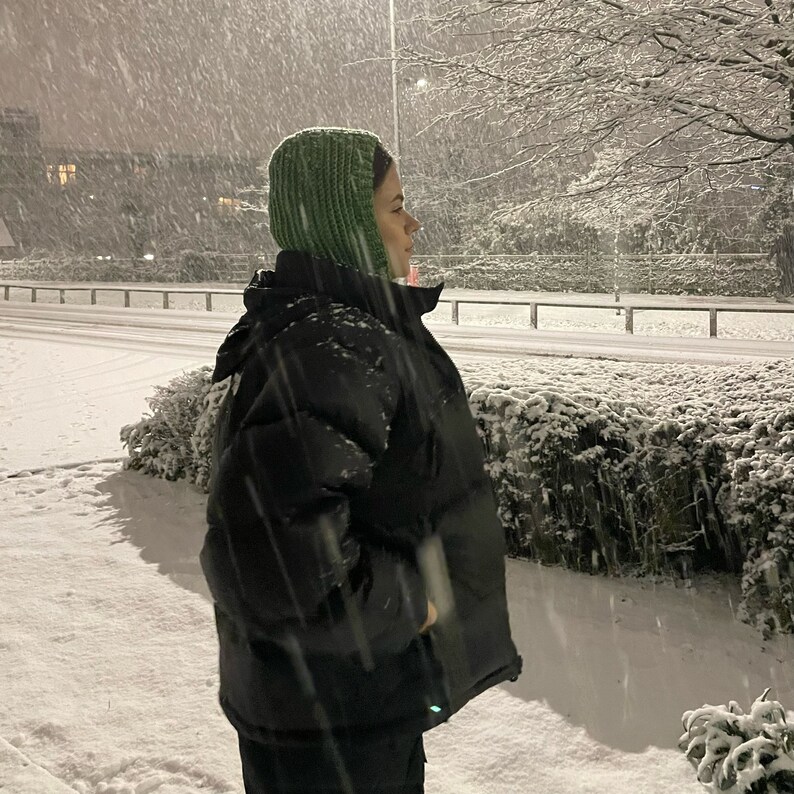 May include: A person wearing a green knitted hat and a black puffer jacket stands in a snowy landscape. The snow is falling heavily and the person is looking off to the side.