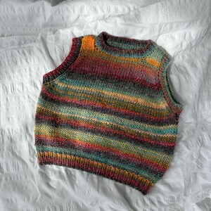 May include: A colorful, hand-knitted vest with horizontal stripes in shades of orange, teal, red, and gray. The sleeveless garment is made of a soft, textured yarn and is displayed on a white surface. The vest has a round neckline.