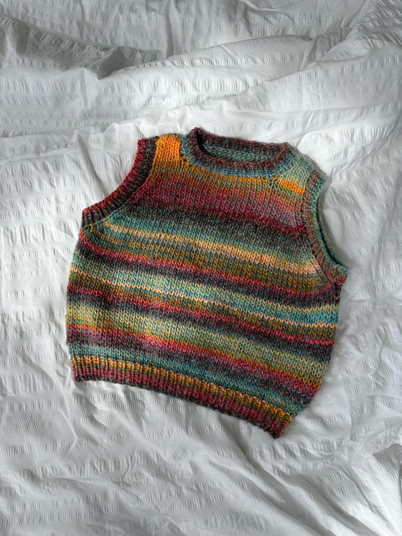 May include: A colorful, hand-knitted vest with horizontal stripes in shades of orange, teal, red, and gray. The sleeveless garment is made of a soft, textured yarn and is displayed on a white surface. The vest has a round neckline.