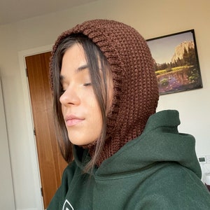 May include: A brown knitted hooded cowl with a long neck covering. The cowl is made of a soft, textured yarn and has a simple, classic design.