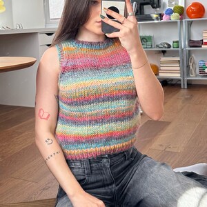 May include: A sleeveless, multi-colored knit top with horizontal stripes in shades of pink, blue, yellow, and green. The top is paired with dark gray jeans. The image is taken indoors, with natural light.