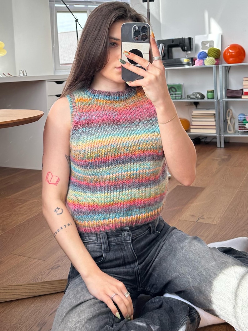 May include: A sleeveless, multi-colored knit top with horizontal stripes in shades of pink, blue, yellow, and green. The top is paired with dark gray jeans. The image is taken indoors, with natural light.