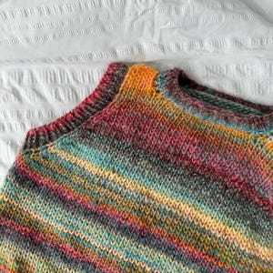 May include: A close-up of a hand-knitted sweater in a vibrant array of colors. The sweater features a mix of pink, orange, yellow, teal, and gray yarns, creating a striped pattern. The neckline and shoulders are visible, showcasing the detailed knit.