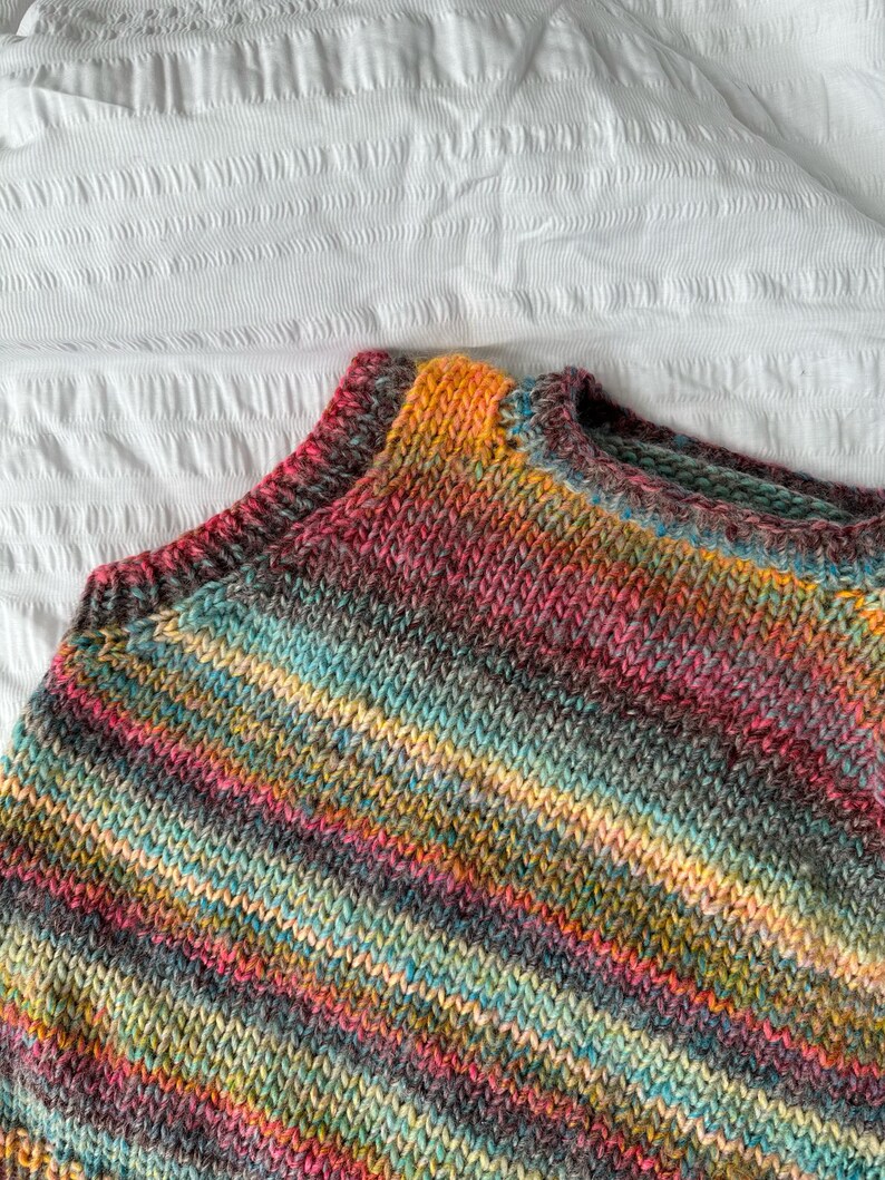 May include: A close-up of a hand-knitted sweater in a vibrant array of colors. The sweater features a mix of pink, orange, yellow, teal, and gray yarns, creating a striped pattern. The neckline and shoulders are visible, showcasing the detailed knit.