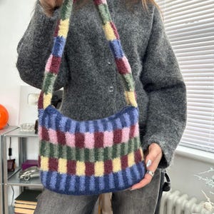 May include: A colorful, knitted shoulder bag with a striped pattern in shades of blue, pink, green, and yellow. The bag has a curved shape and a matching strap. The bag is made of a soft, textured material.
