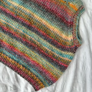 May include: A sleeveless, hand-knitted top in a vibrant array of colors including pink, teal, yellow, and gray. The yarn is arranged in horizontal stripes, creating a textured, cozy appearance. The garment is displayed on a white surface.