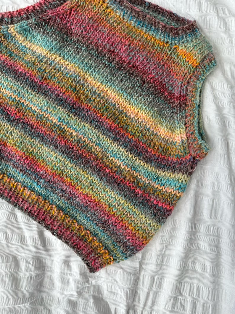 May include: A sleeveless, hand-knitted top in a vibrant array of colors including pink, teal, yellow, and gray. The yarn is arranged in horizontal stripes, creating a textured, cozy appearance. The garment is displayed on a white surface.