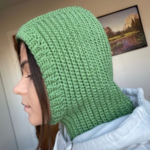 May include: A green crocheted hooded cowl with a ribbed texture. The cowl is worn by a person with long dark hair.