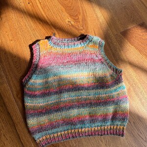 May include: A colorful, hand-knitted vest with a round neckline. The vest features horizontal stripes in shades of pink, blue, yellow, and purple. The garment is made of soft yarn and is suitable for layering. The vest is displayed on a wooden surface.