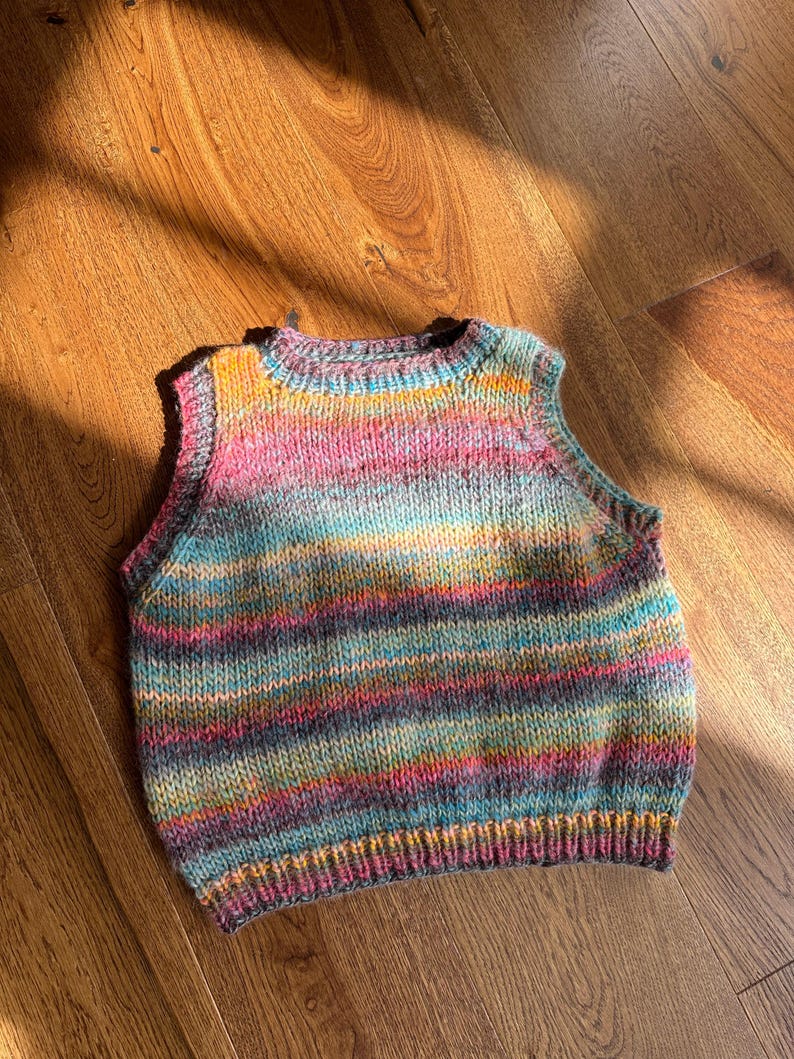 May include: A colorful, hand-knitted vest with a round neckline. The vest features horizontal stripes in shades of pink, blue, yellow, and purple. The garment is made of soft yarn and is suitable for layering. The vest is displayed on a wooden surface.