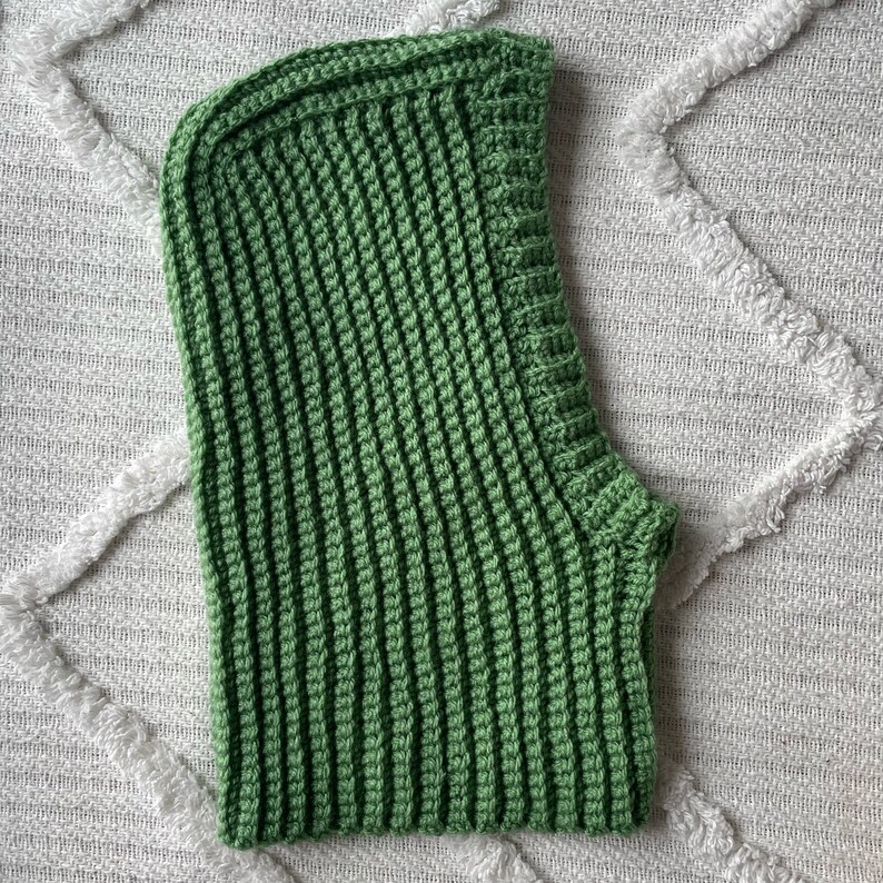 May include: A green crocheted hooded scarf. The scarf is made with a simple stitch and has a long, flowing shape.