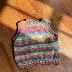 May include: A hand-knitted, sleeveless vest in a vibrant array of colors including pink, blue, yellow, and purple. The vest features horizontal stripes and a ribbed hem and neckline. The garment is made of soft yarn.