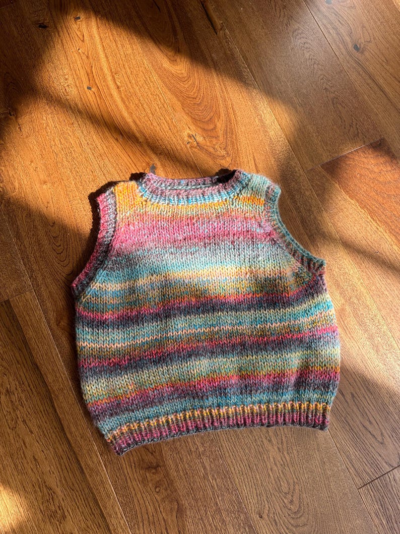 May include: A hand-knitted, sleeveless vest in a vibrant array of colors including pink, blue, yellow, and purple. The vest features horizontal stripes and a ribbed hem and neckline. The garment is made of soft yarn.