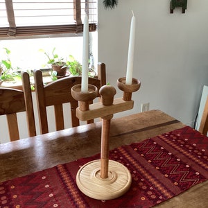 May include: A wooden candle holder with two white candles. The holder has a round base and a tall, slender stem. The candles are in a natural wood finish.