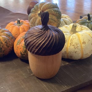 May include: A wooden acorn-shaped container with a dark brown, carved top and a light brown base. The container is surrounded by various pumpkins in shades of orange, green, and white, arranged on a wooden surface.