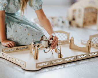 May include: A wooden toy horse jumping course with a toy horse and rider. The course is made of light-colored wood and includes fences and a rectangular base. The horse is white with blue accents, and the rider wears a red jacket.