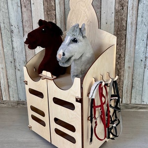 May include: Wooden horse stable playset featuring two plush horse heads, a wooden horse head cutout, and hooks for hanging reins. The stable has a door with four slots and is made of light wood.