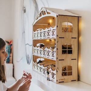 May include: A wooden toy stable with four levels, each with a stall for a horse. The stable has a light brown finish and a white roof. A small figure of a person is standing in front of the stable. The text "in Timber Q" is printed on the bottom right corner of the stable.